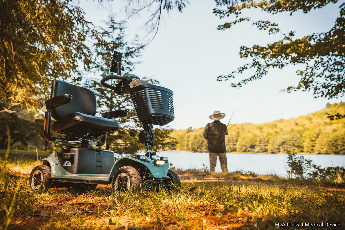 Sage Baja Bandit mobility scooter parked on a grassy lakeshore beneath trees, with a person fishing beside the water in the background.