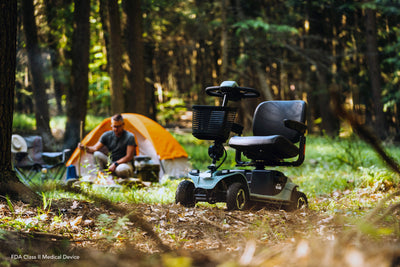 Sage Baja Bandit mobility scooter parked at a wooded campsite, with an orange tent and a person tending a campfire in the background.