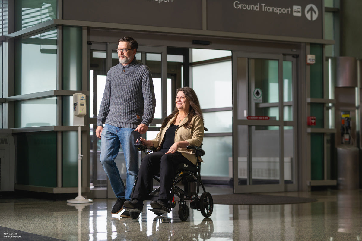 User driving the Jazzy Carbon 27X through an airport with a companion, highlighting travel-friendly mobility.
