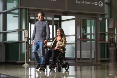 User driving the Jazzy Carbon 27X through an airport with a companion, highlighting travel-friendly mobility.