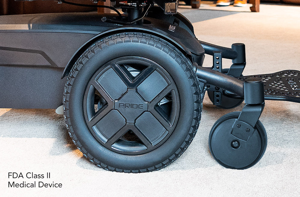 Close-up of a black Pride Jazzy EVO powerchair’s treaded drive wheel, front caster, and footplate, with text reading “FDA Class II Medical Device.”
