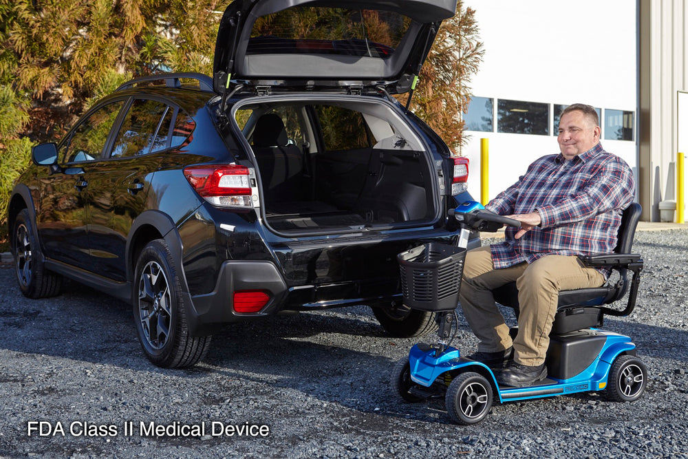 Man seated on a blue Pride Revo 2.0 scooter beside a black SUV with its rear hatch open, on a gravel parking area.