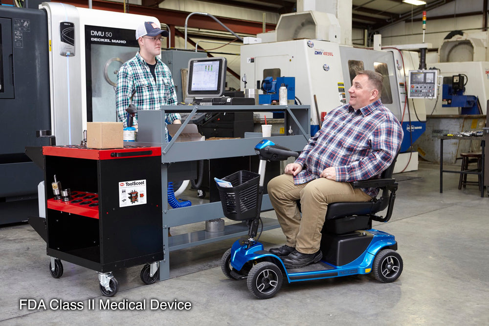 Man seated on a blue Pride Revo 2.0 scooter talking with another man beside a tool cart in a machine workshop.