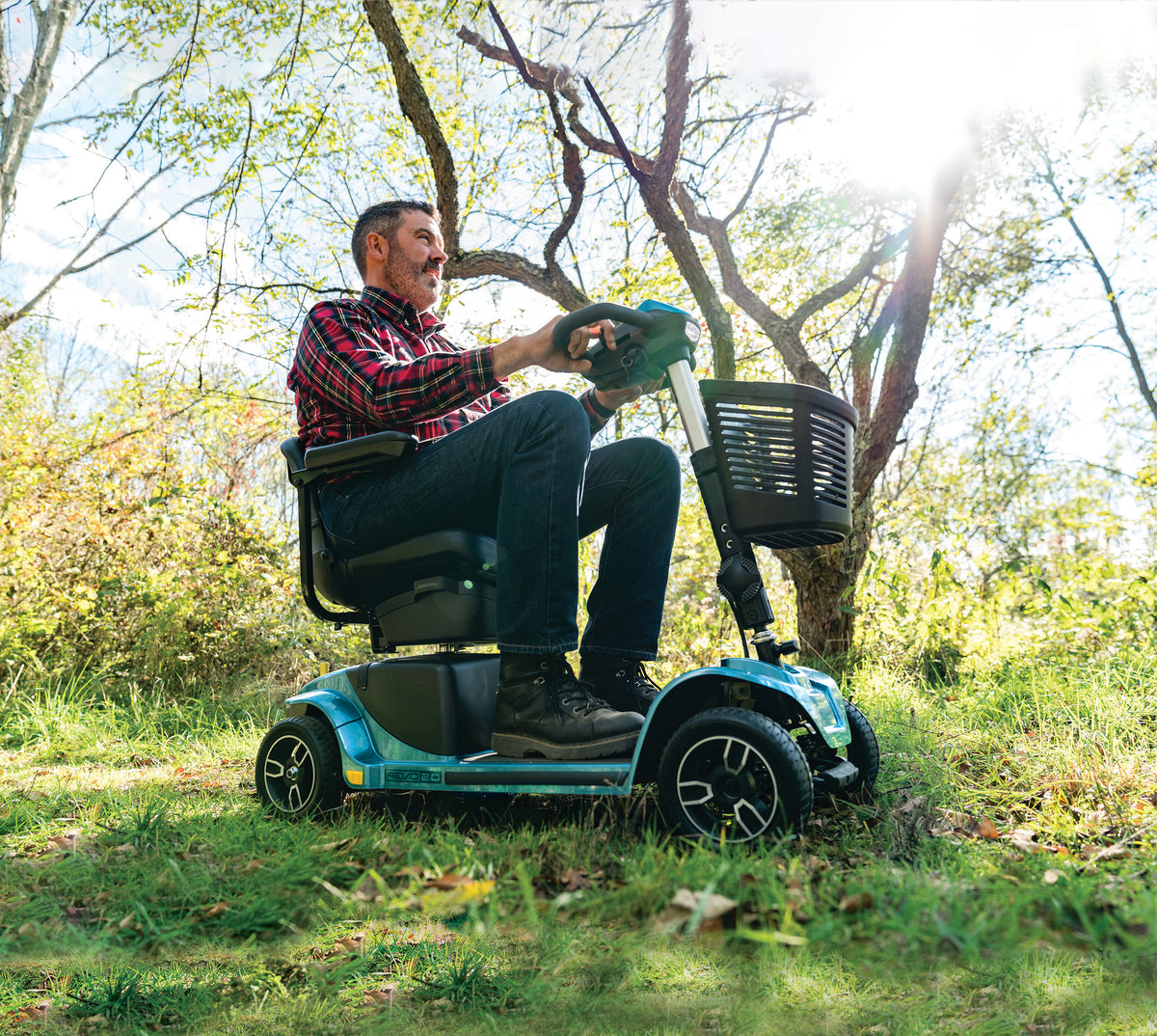Side view of a man riding a blue Pride Revo 2.0 four-wheel mobility scooter on grass beneath sunlit trees.